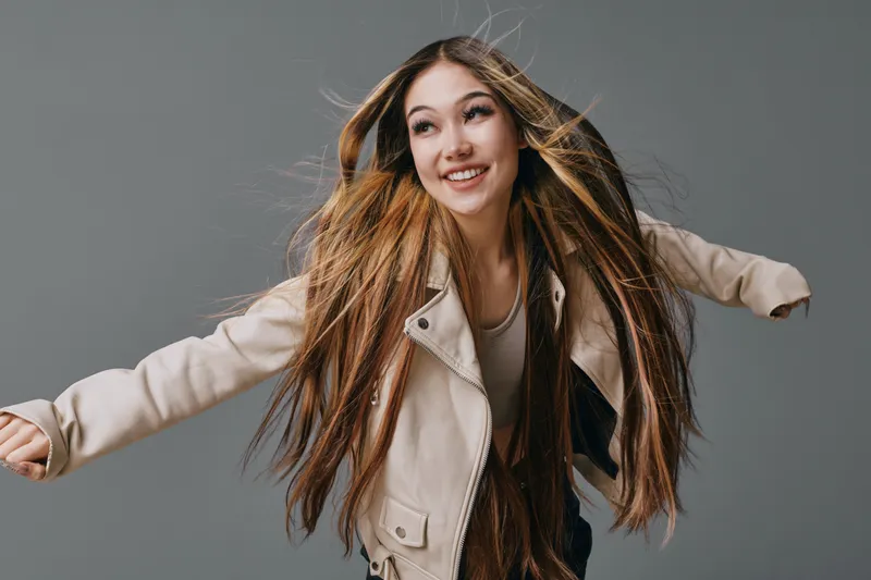 Young woman with long hair smiling and posing in beige jacket on gray background for fashion, beauty and lifestyle studio portrait with casual style and joyful expression.