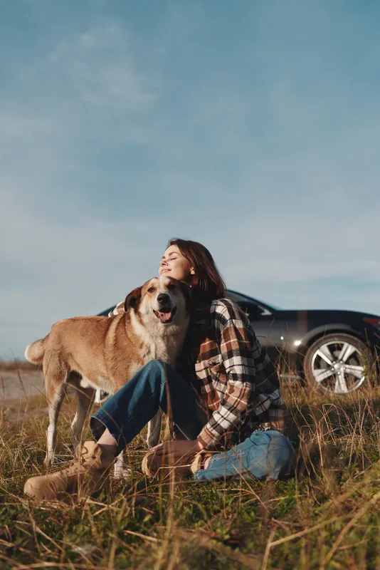 Woman sits with dog in a grassy field beside a sleek car, enjoying sunny weather and nature during a relaxed outdoor moment of companionship and joy together.