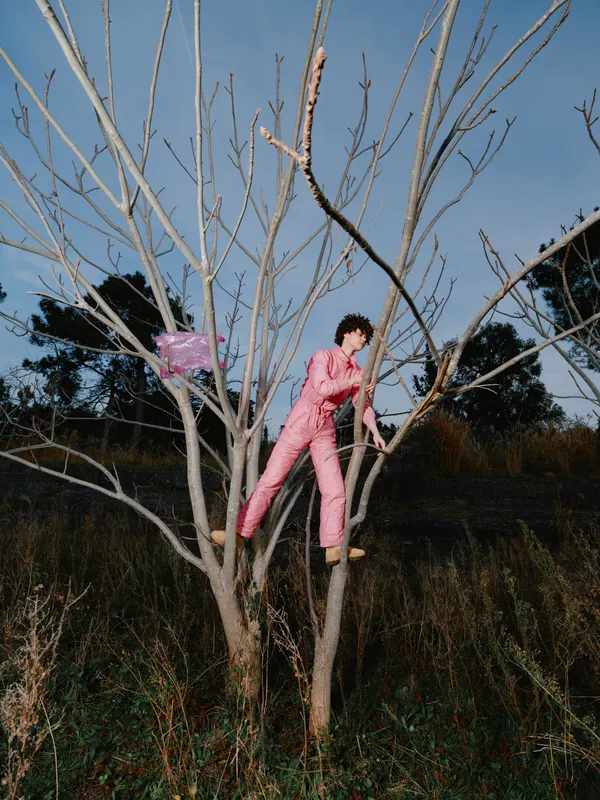 Person in pink jumpsuit climbs a leafless tree in an open field, balancing on branches for adventure and exploration against a bright blue sky, highlighting outdoor activity, bold color contrast and