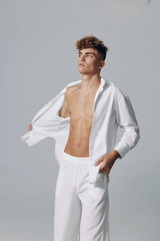 Young man with curly hair posing in white open shirt and pants, showing toned abs and confident expression against plain gray background in studio.