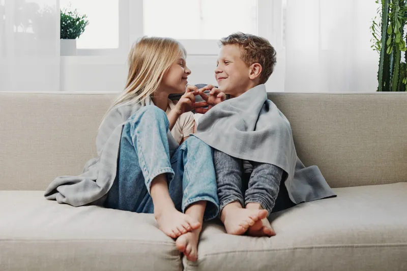 Happy siblings wrapped in cozy blanket sitting barefoot on sofa smiling and playing together in bright living room.