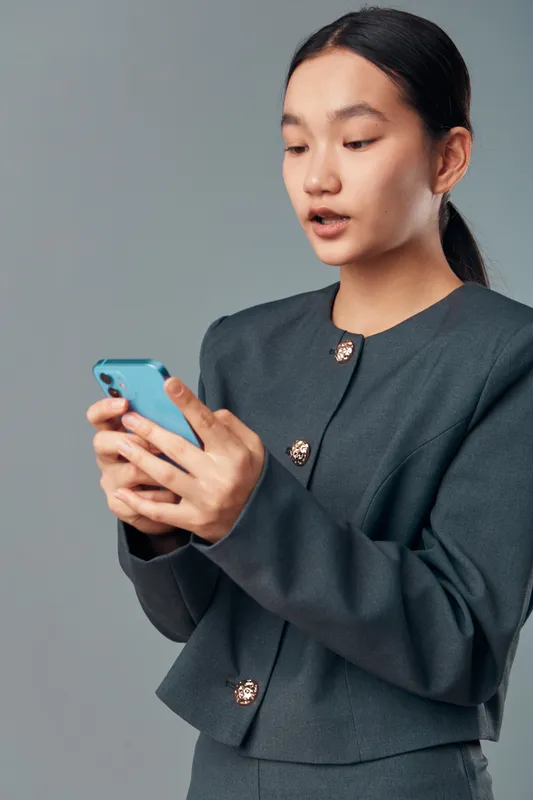 Young woman in business attire looking at smartphone screen with focused expression isolated on grey background. Professional female holding phone for communication or reading messages.