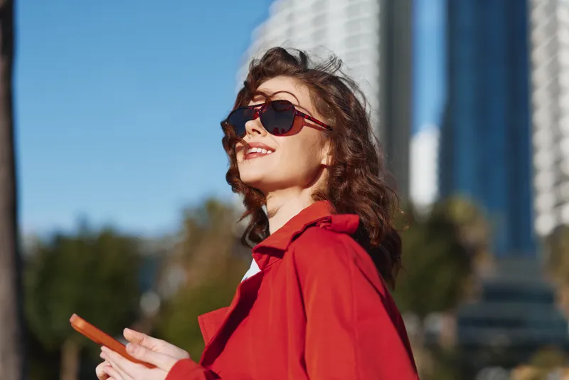 Young woman wearing red coat and sunglasses smiling outdoors in sunny urban environment holding smartphone enjoying bright day with clear blue sky and blurred buildings background