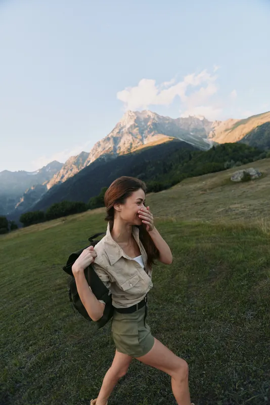 Young woman with backpack hikes across a grassy mountainside, enjoying a sunny day and fresh air, in casual utility outfit as mountains loom in the background.