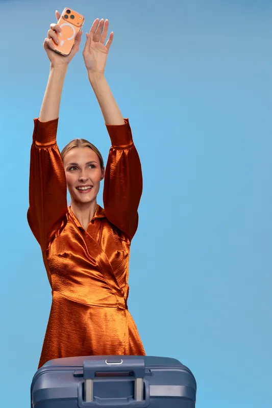 Smiling woman in orange satin dress holds mobile phone up and stretches arms, standing behind blue suitcase on blue background. Concept of travel, vacation, happiness, and modern lifestyle.