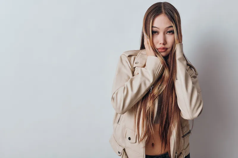 Young woman with long straight hair wearing beige jacket posing with hands near face on gray background isolated. Fashion, beauty and style concept for model portrait.