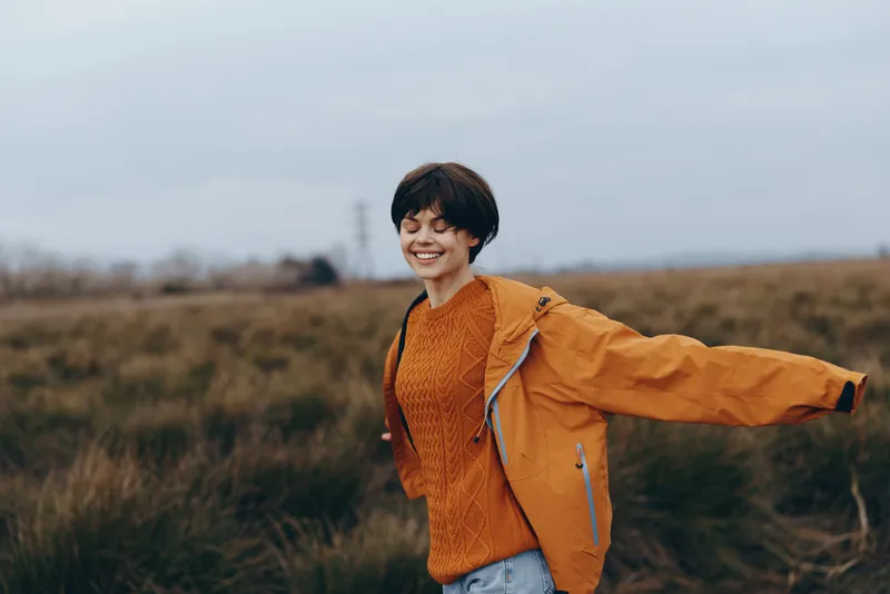 Outdoor smile of young person in orange jacket, standing in field, nature landscape under cloudy sky, soft daylight, casual lifestyle moment, friendly youth portrait, for outdoor travel