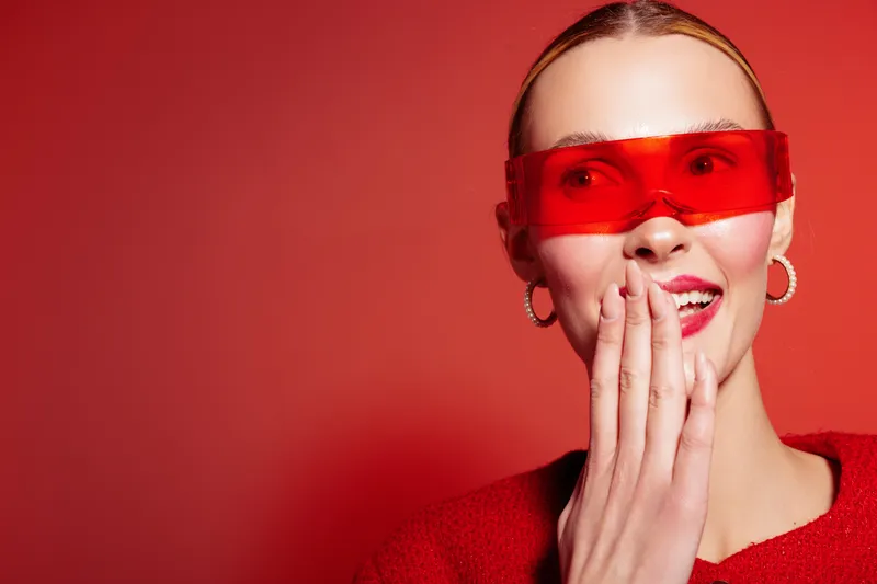 Smiling woman in red sweater and red transparent glasses covers mouth with hand, looking aside. Bright red background creates vibrant and playful mood, concept of fashion and happiness.