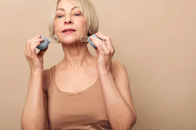 Mature woman with short blonde hair uses facial massage rollers on both cheeks against beige background. Concept of anti aging skincare, self care, wellness, beauty routine, and healthy lifestyle.