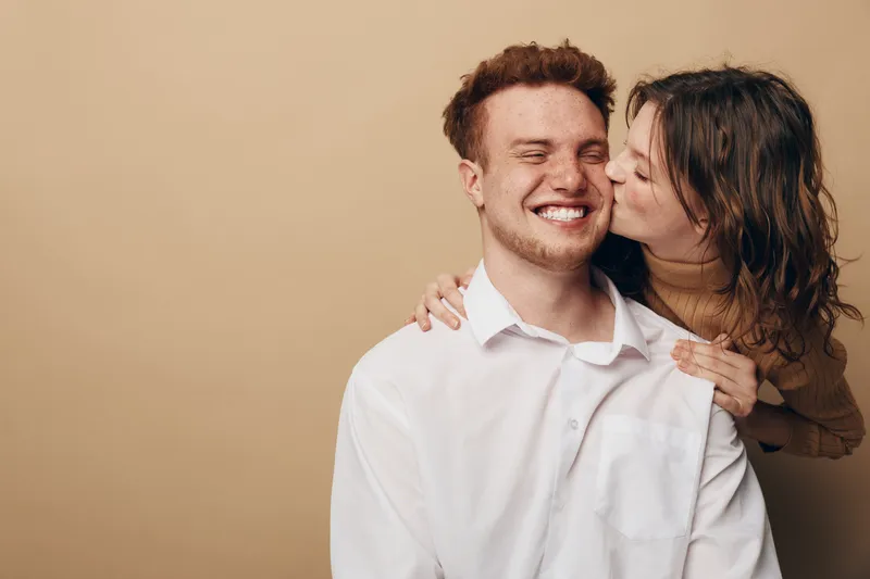 Happy young couple in casual clothes share affectionate moment, woman kissing man on cheek. Warm beige background, natural light, concept of love, relationship, intimacy, and happiness.