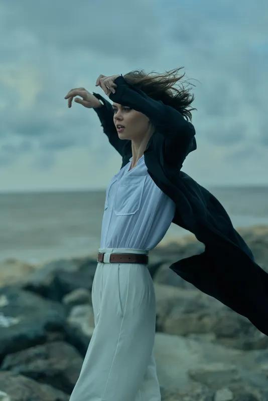 Woman in white dress and dark coat on a windy beach, catching the sea breeze as waves crash, motion and fashion captured for outdoor photography, dramatic natural setting with an energetic pose by