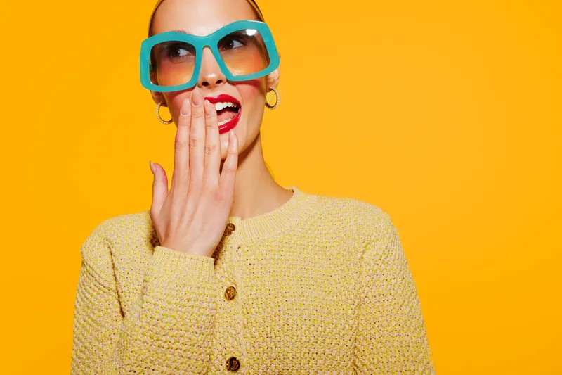 Stylish woman in yellow sweater and large turquoise glasses covers mouth with hand, expressing surprise or amusement. Bright yellow background creates vibrant and cheerful mood.