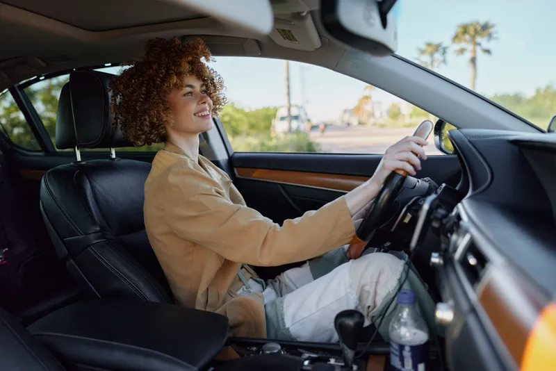 Smiling woman with curly hair driving a car, wearing a beige jacket. Close up on interior with natural light. Concept of lifestyle, travel, and self expression. Retro film color adds warmth.