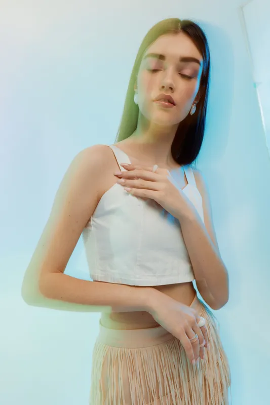Young woman with long hair poses with closed eyes and hand on chest wearing a white top and beige skirt. Soft lighting creates a serene mood. Concept of beauty fashion and elegance.