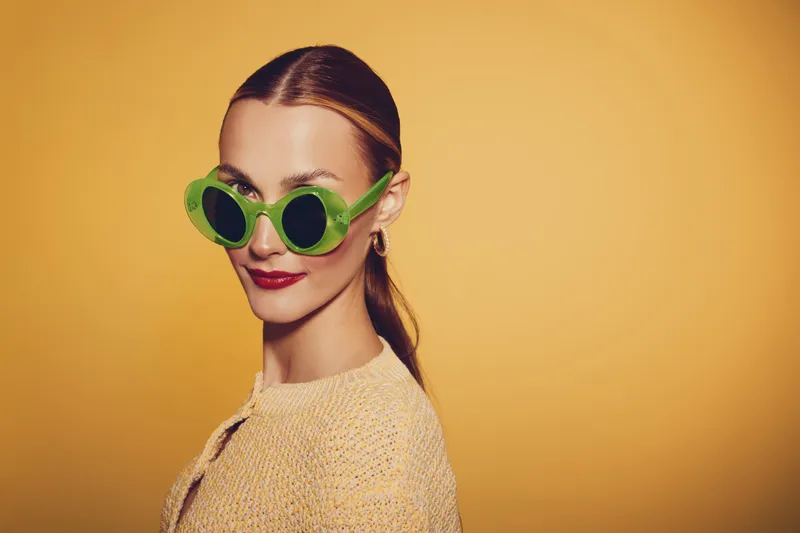 Stylish woman with green sunglasses and red lipstick poses against yellow background. She wears a beige knitted sweater and has slicked-back hair. Concept of fashion and retro style.