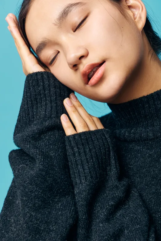 Woman with closed eyes and relaxed face wearing dark sweater posing with hand on cheek against blue background. Portrait of calm peaceful female with smooth skin and natural expression.