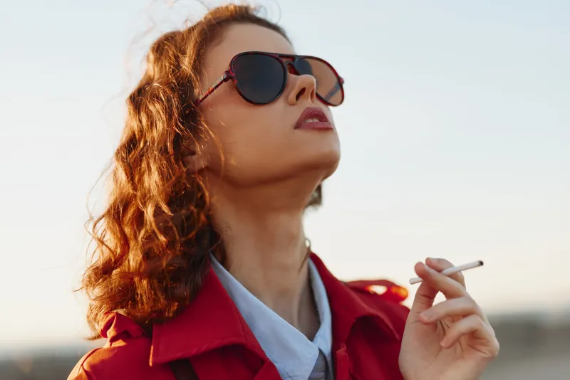 Stylish redhaired woman wearing sunglasses and a red coat smokes a cigarette, gazing upward against a clear blue sky under warm sunlight on a calm day.