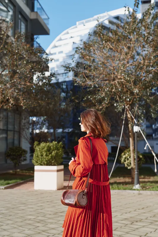One young woman in a red pleated dress stands in a sunny urban plaza, turned to the side with a brown leather crossbody bag, conveying contemporary street style and confidence.