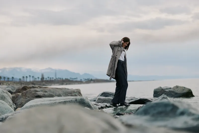 Young woman in coat stands on rocky shore near ocean, looking down with hand on head. Overcast sky and distant mountains create calm and reflective mood. Concept of solitude and nature.