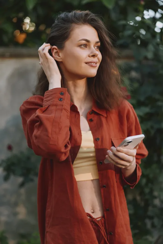 A woman in a rust jacket enjoys outdoor lifestyle, holding a smartphone, smiling softly, looking to the side. A casual portrait of fashion and relaxed mood