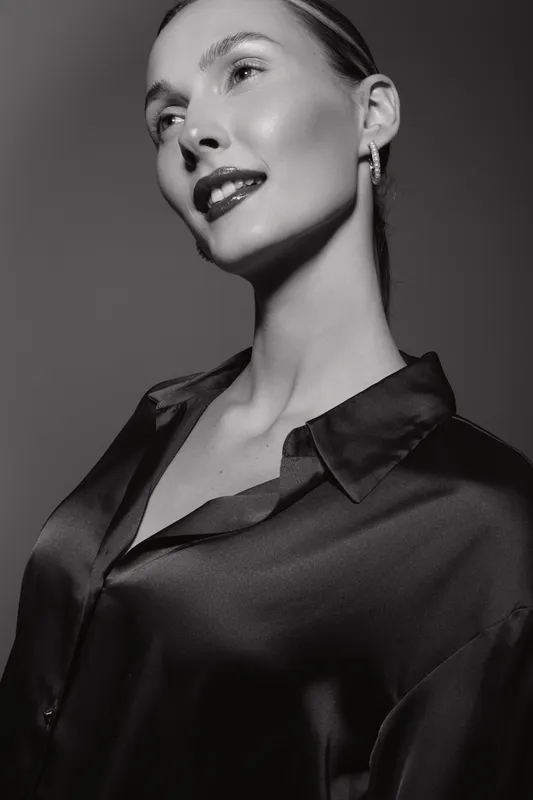 Elegant white woman with light skin wears satin blouse and earrings, posing in black and white studio portrait.
