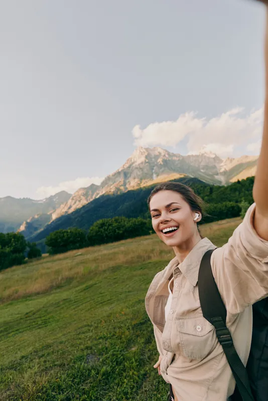 A smiling woman poses outdoors on a hillside, taking a selfie with a backpack visible, amid bright mountains and blue sky, conveying an active lifestyle moment.