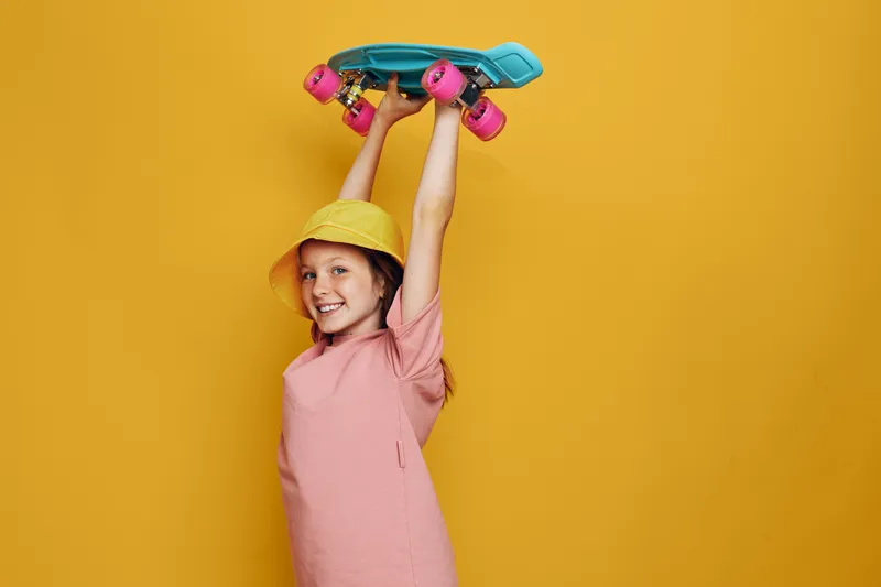 Smiling girl in a yellow hat holds a blue skateboard above her head. She wears a pink t-shirt and stands against a solid yellow background. Concept of childhood, fun, and sport.