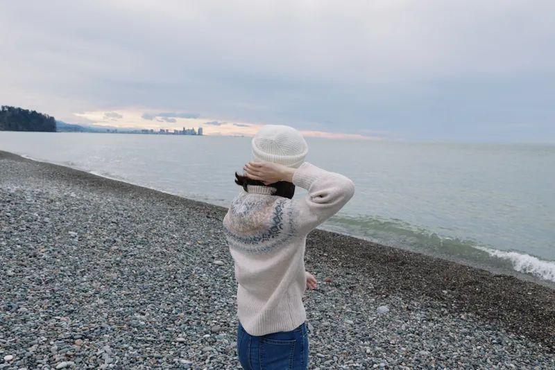 woman on a pebble beach facing the sea in a white sweater and knit hat, gazing toward the horizon as a distant skyline appears under a calm, cloudy sky