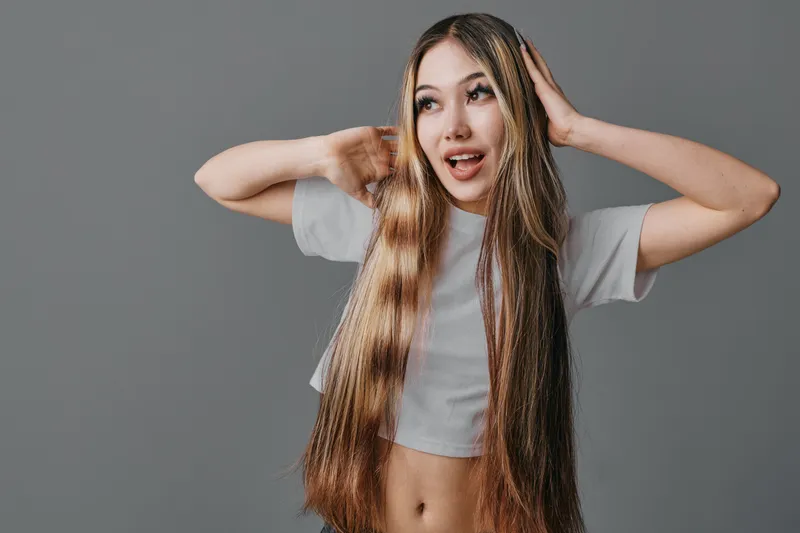 Woman with long straight hair posing in white crop top on gray background, expressing surprise with hands near head and open mouth, casual style and natural look indoors.