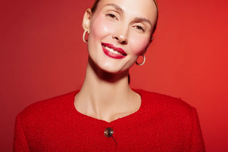 Smiling woman with red lipstick and hoop earrings poses against red background wearing red textured jacket. Concept of beauty, fashion, confidence, and vibrant style in studio lighting.