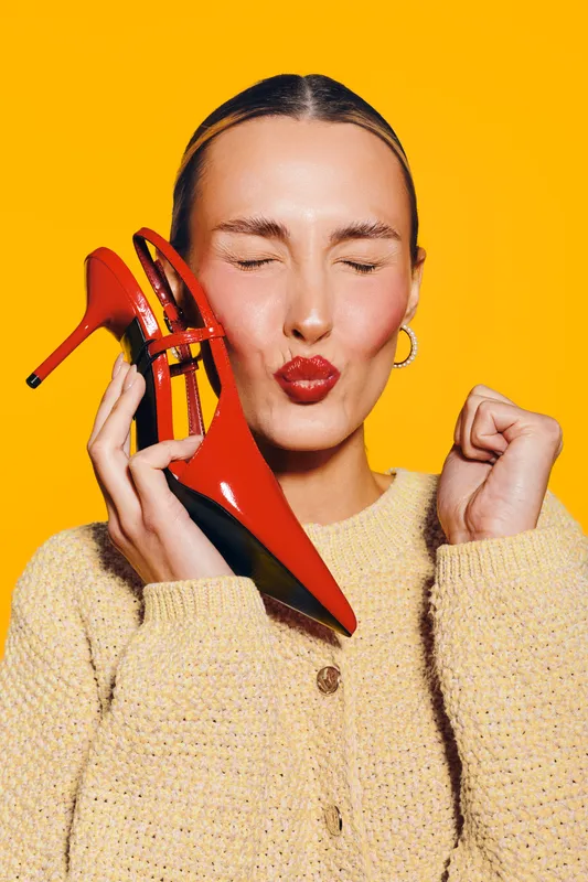 Young woman with closed eyes holds red high heel shoe near face, wearing beige sweater on yellow background. Concept of fashion, style, and happiness with bright colors.