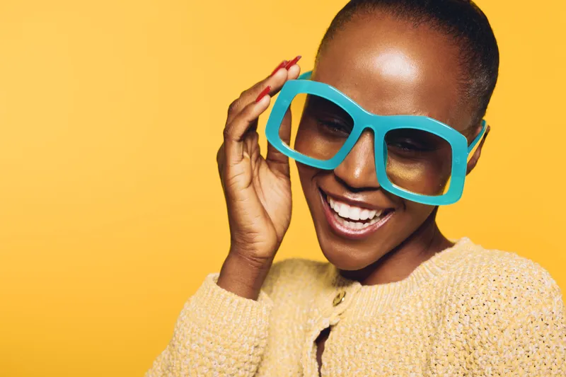 Happy African woman with dark skin wearing large blue sunglasses and a yellow sweater smiles brightly against a vibrant yellow background. Concept of joy, fashion, and summer style.