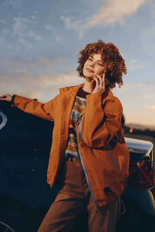 Woman lifestyle outdoors at sunset, curly hair, relaxed pose, orange jacket and scarf, touching chin near a parked car, warm evening light, soft sky