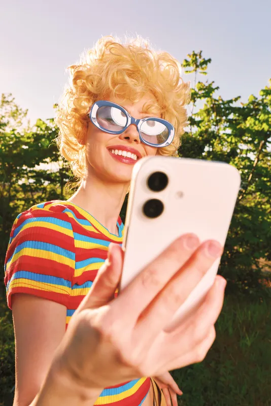 Smiling woman with curly hair taking a selfie outdoors, wearing a striped shirt and retro sunglasses. Bright sunlight creates a vibrant, carefree lifestyle mood, warm tones and playful energy.