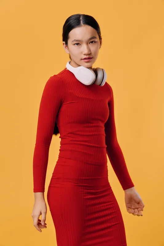 Young woman in red long sleeve dress wearing white headphones around neck standing against orange background looking at camera with serious expression in studio portrait.