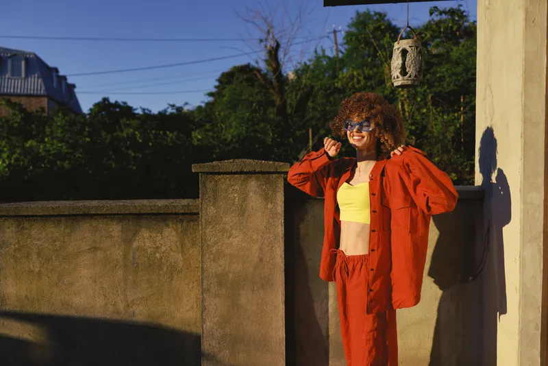 Stylish woman with curly hair poses confidently on a sunny terrace, wearing an orange suit and yellow crop top, leaning against a warm wall with sunglasses and a carefree vibe