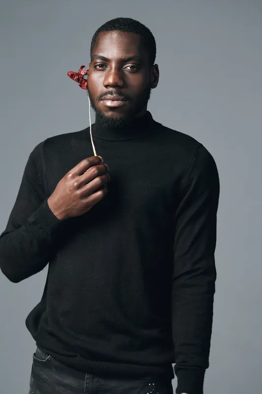 African man in black sweater holding red flower near face in creative conceptual visual style. Portrait with serious expression on gray background for artistic photography series.