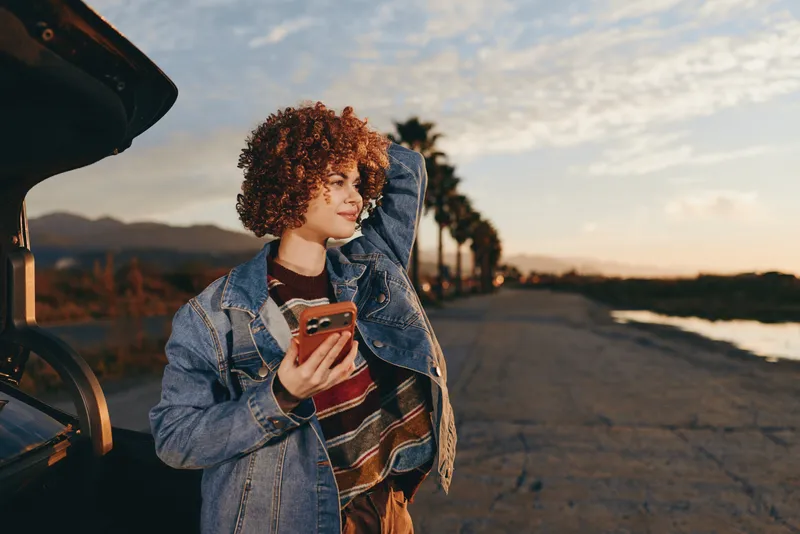 Woman lifestyle outdoors at sunset, curly hair and denim jacket, smiling with smartphone in hand, leaning by open car hood, calm road, distant mountains, golden sky, natural light