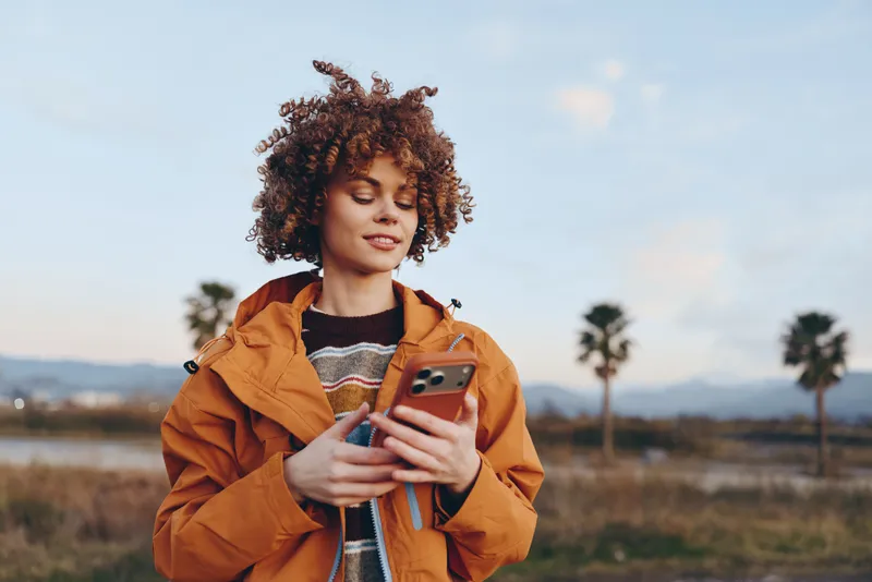 Woman lifestyle outdoors portrait, curly hair, wearing orange jacket, holding smartphone, looking at screen, relaxed active smile, palm landscape in distance, daytime field, bright sky