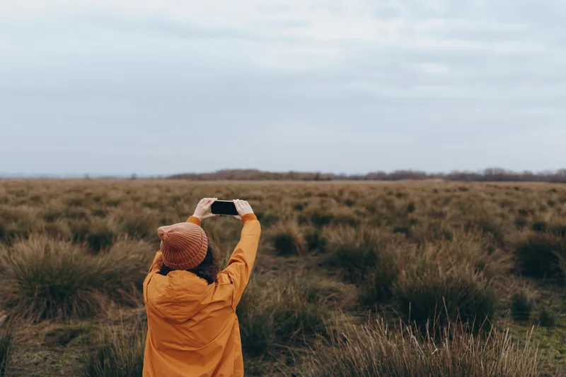 Woman outdoor nature field landscape lifestyle cloudy grass hat jacket, in tall grass, holding binoculars and watching distant horizon under overcast clouds, calm leisure moment