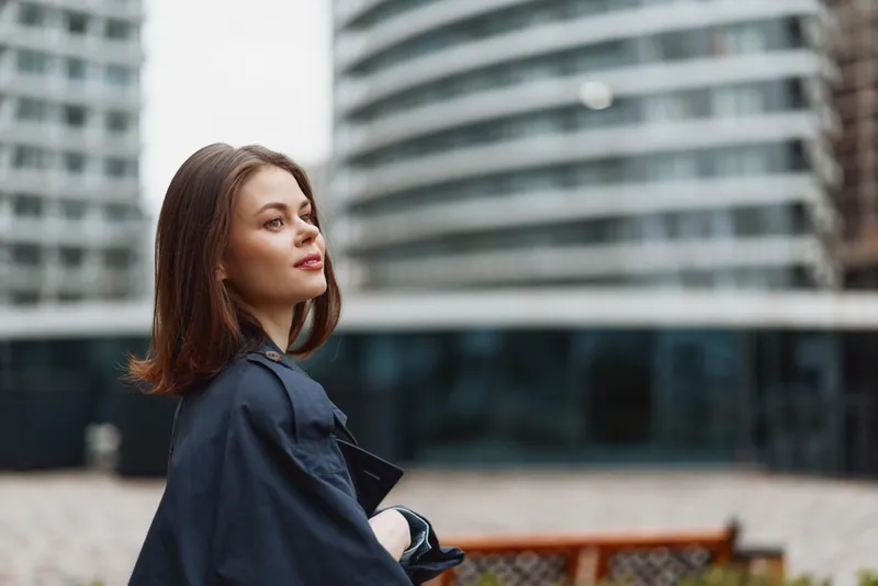 Urban portrait of a young woman in a navy coat standing beside a bench with modern buildings behind, exuding calm confidence and a subtle, reflective smile.