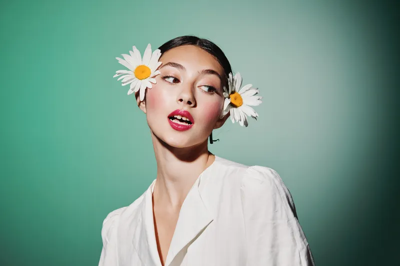 Young woman with creative makeup and two flower decorations in hair poses expressing elegance and youth on a smooth green background. Concept beauty fashion art.