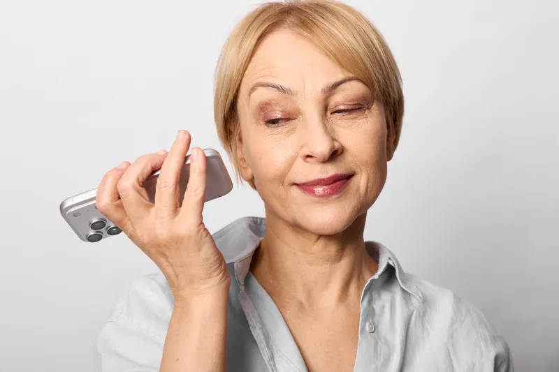 Woman holding smartphone for a call, phone near ear, relaxed eyes, senior person in soft studio light, gray background, calm communication concept, everyday technology, wellbeing, gentle smile