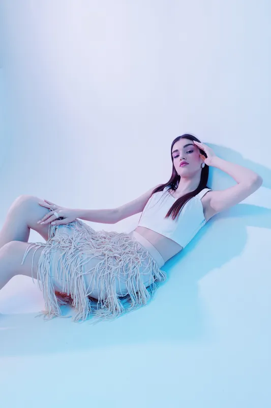 Young woman in a stylish outfit poses reclining on the floor in a bright studio. The image highlights beauty, fashion, and modern style with soft lighting and a calm mood.