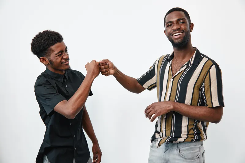 Two young black men with dark skin smiling and giving fist bump in casual clothes on white background. Friendship, happiness and positive gesture between male friends.