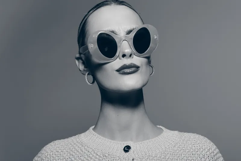 Elegant white woman with pale skin wearing oversized round sunglasses and knitted top poses confidently in studio. Concept for fashion, style, and modern accessories.