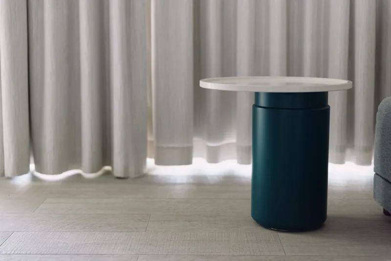 Round table on a blue pedestal base stands in a minimal living room with light curtains and soft flooring, showcasing modern interior design.