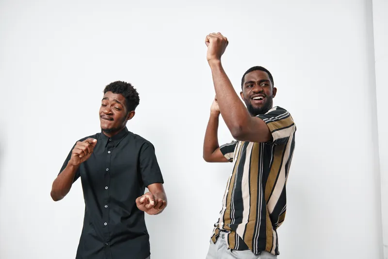 Two African men with dark skin dancing and enjoying music together in casual clothes on white background. Friends having fun and expressing happiness indoors.