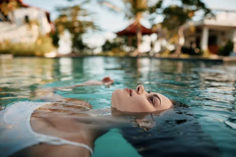 Relaxed woman floating on water surface in outdoor pool, eyes closed, peaceful expression, natural light, summer day, calm atmosphere, serene lifestyle moment