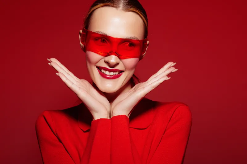 Smiling woman in red sweater and red sunglasses poses with hands near face on red background. Concept of fashion, style, happiness, and vibrant color contrast in studio portrait.